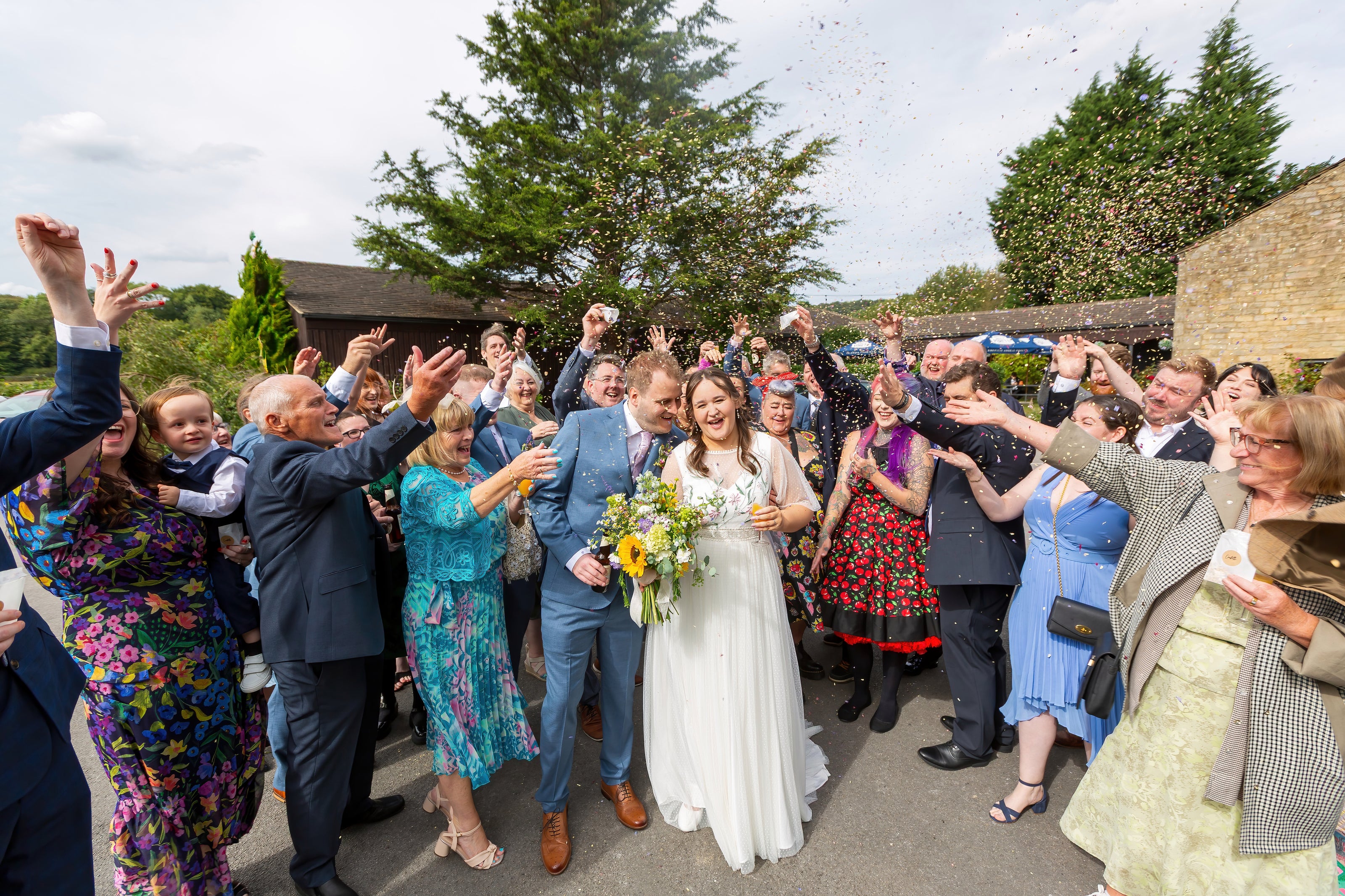 Wedding celebration with the bride and groom (Helen & Dan) surrounded by guests outdoors, throwing confetti in the air