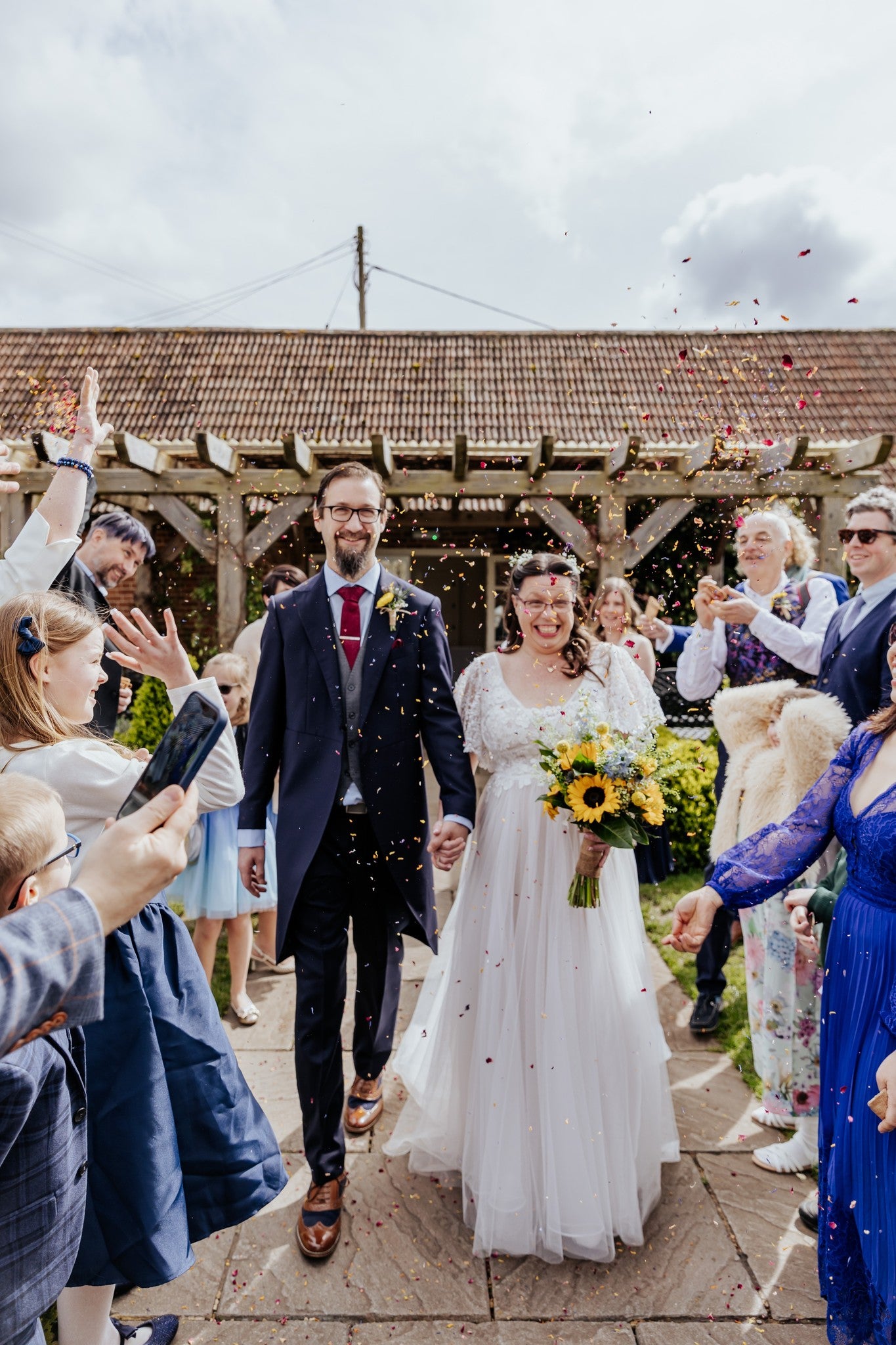 Wedding couple walking down the aisle with guests on a cloudy day during the confetti toss. The bride carries a sunflower bridal bouquet.
