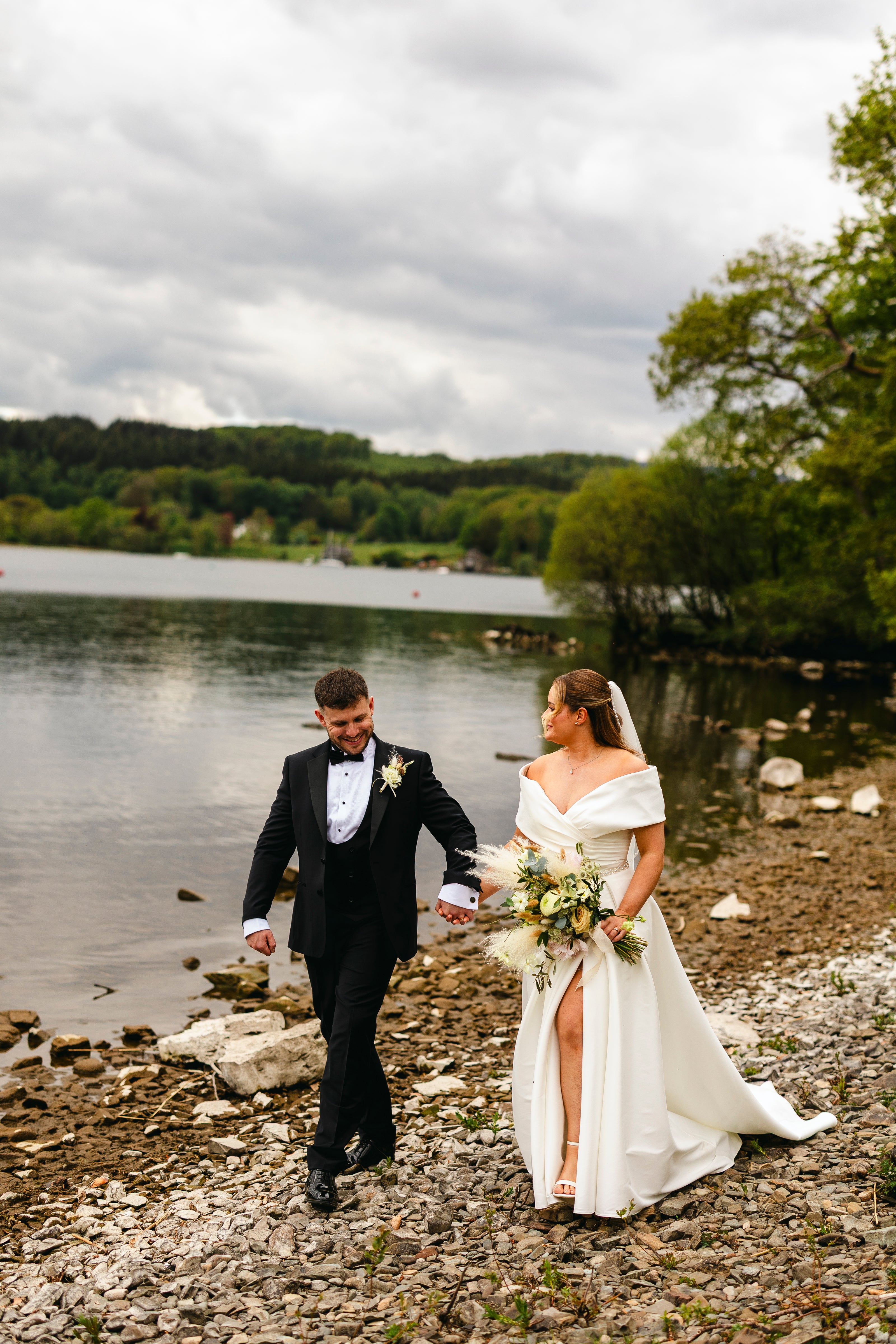Freshly married couple walking by a lake on a cloudy day, holding hands. The bride in her dress holds a fluffy bridal bouquet.