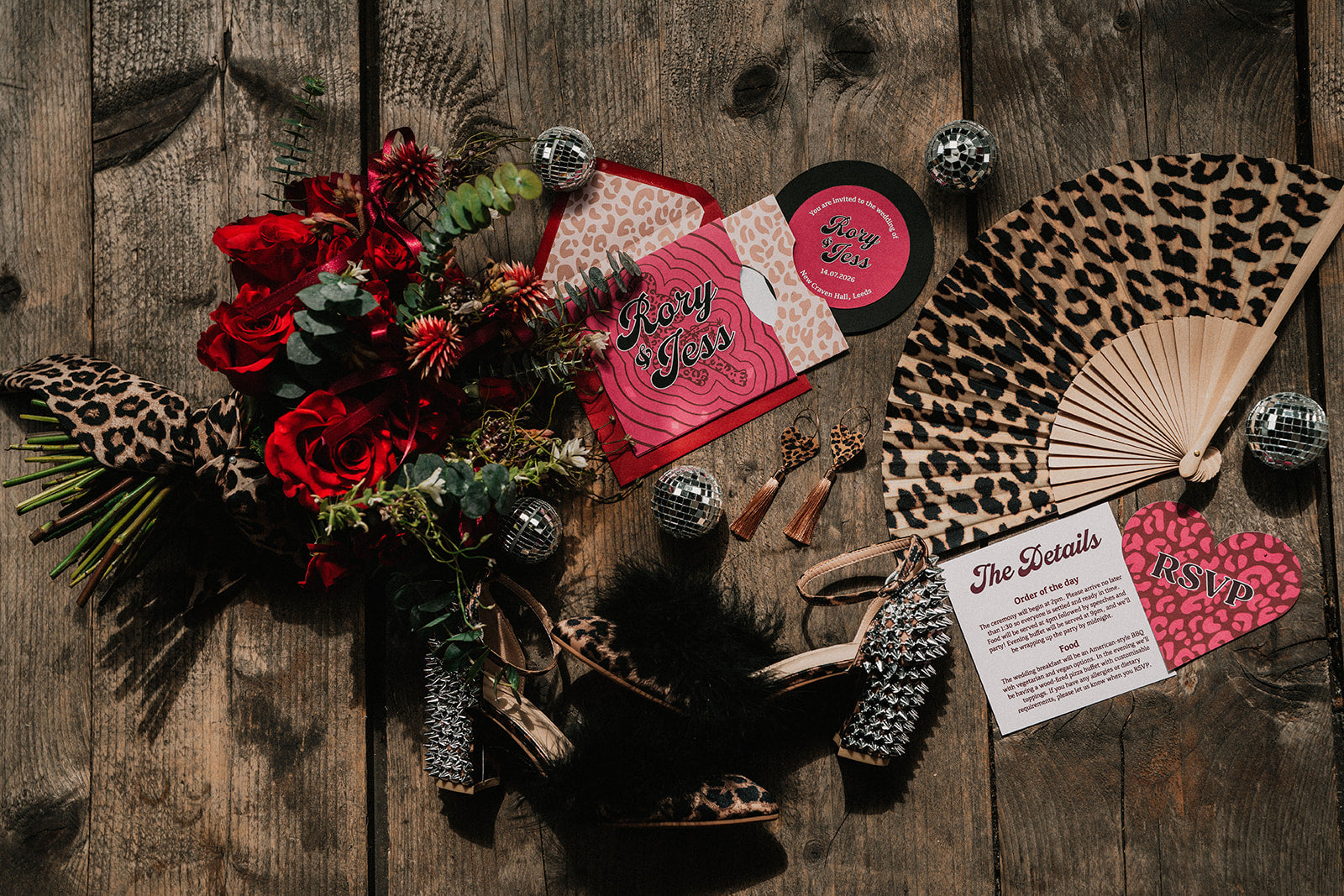 A styled flat-lay of a gothic leopard-print wedding invitation suite on a rustic wooden background, including a main invitation card, RSVP card, envelope, matching leopard-print napkin, fan, feather, and red rose bouquet.

Photography by CRT Weddings