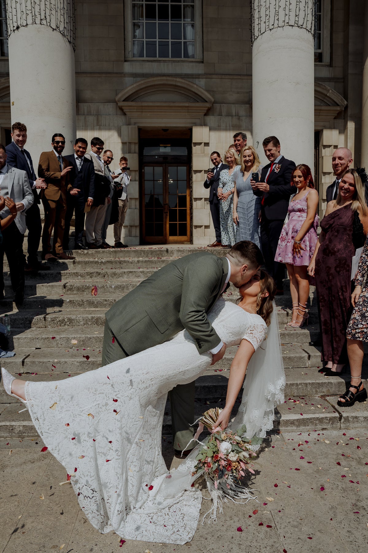 Wedding scene with a couple on the steps of a building, surrounded by guests after the confetti toss. The groom has the bride dipped in a kiss. 
