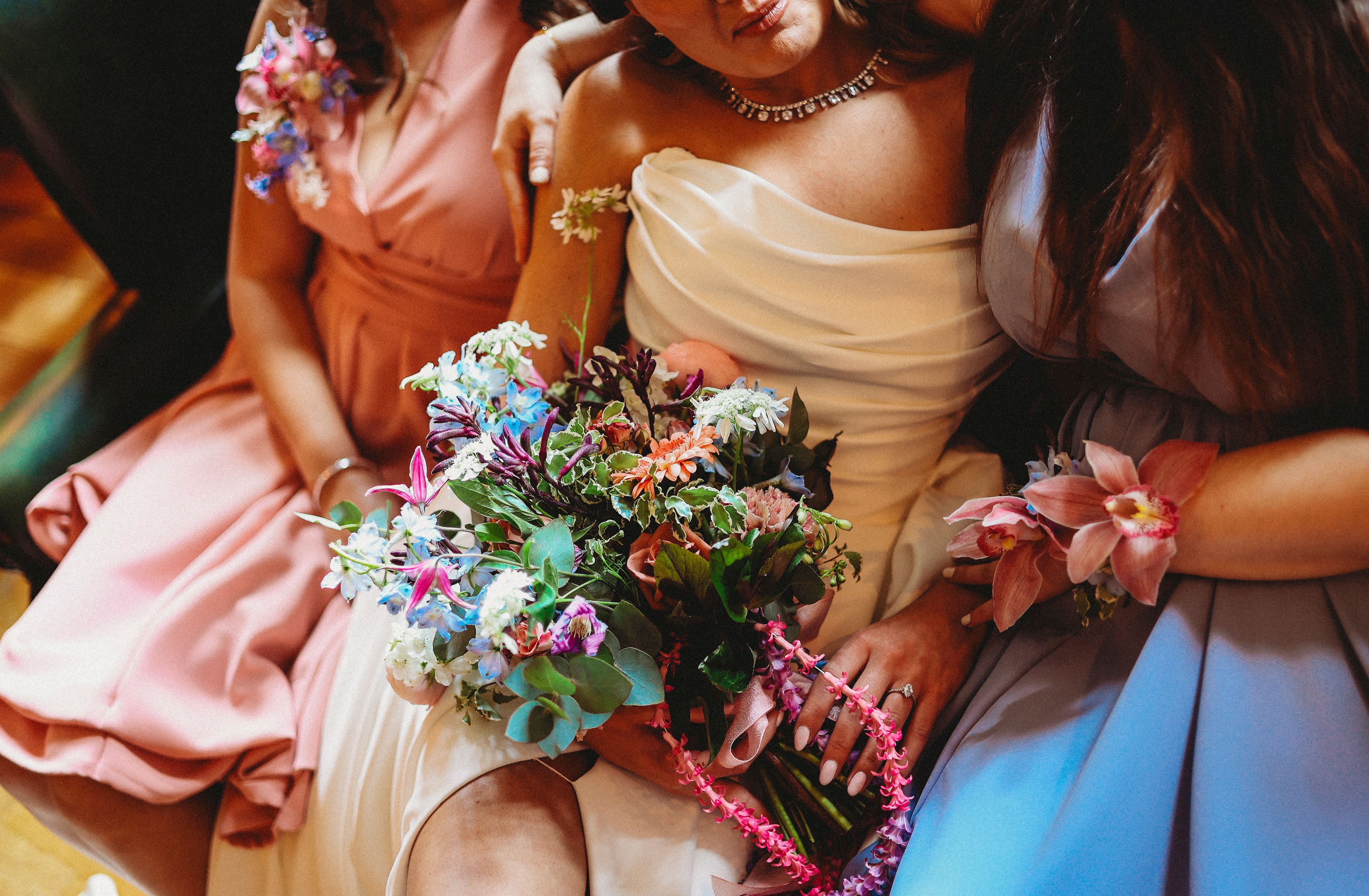 Three women in formal dresses holding a bouquet of flowers. An orchid corsage is worn on the wrist of the woman in the blue dress, the bride holds a bridal bouquet.
