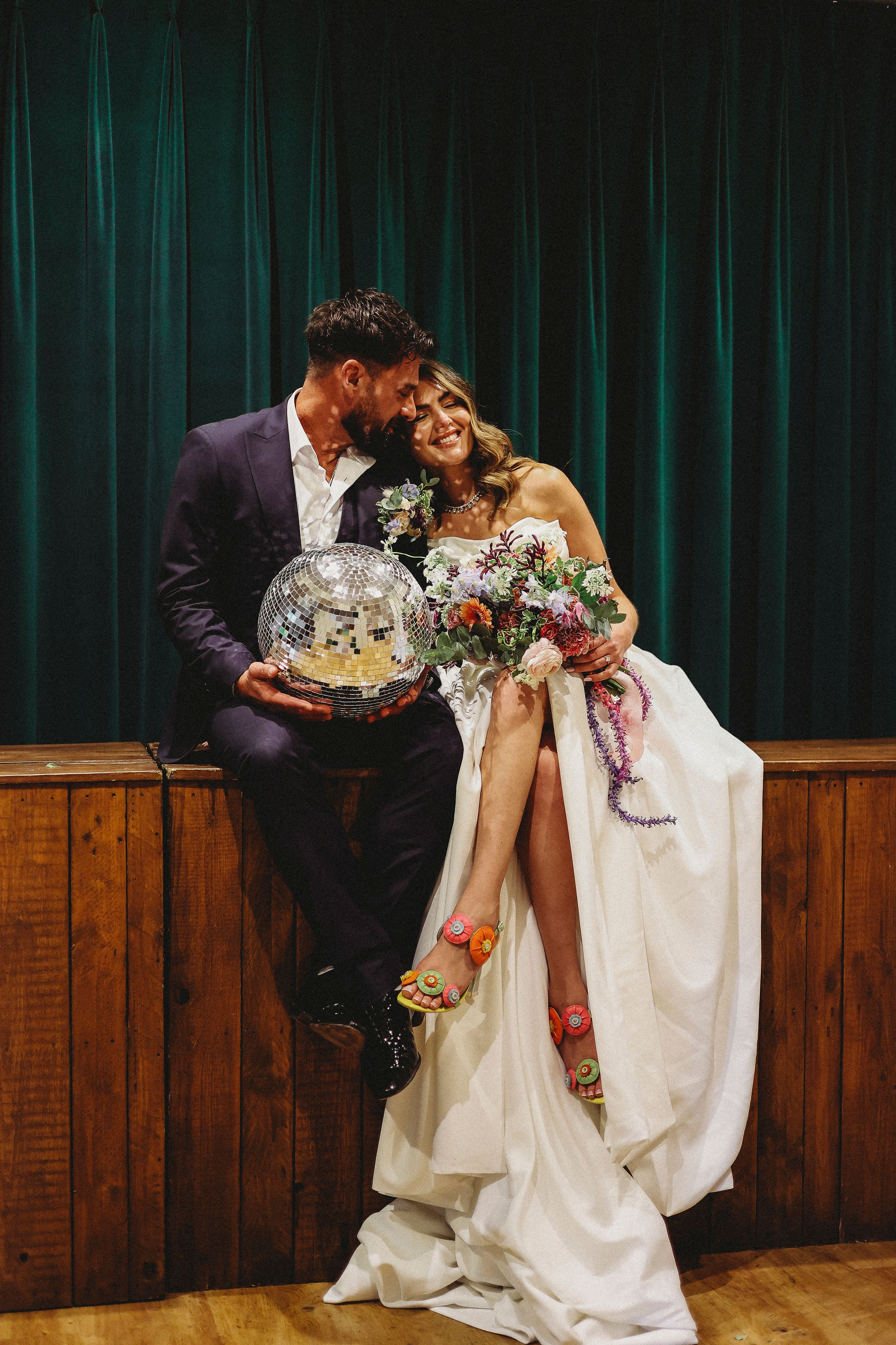 Bride and groom holding a disco ball and floral bridal bouquet against a green curtain background.