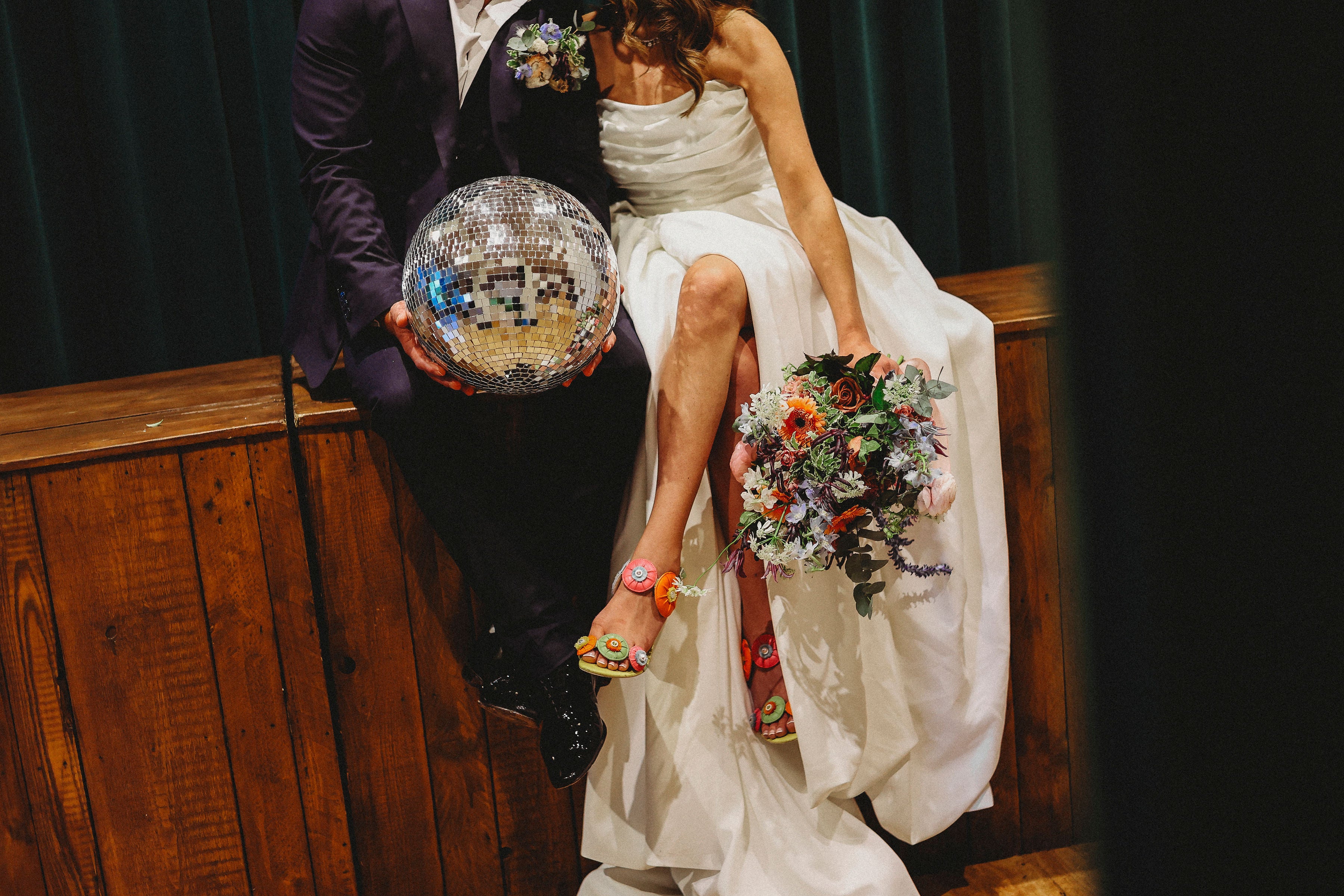 Groom in a suit holding a disco ball next to the bride in a wedding dress with a bouquet, sitting on a wooden bench.
