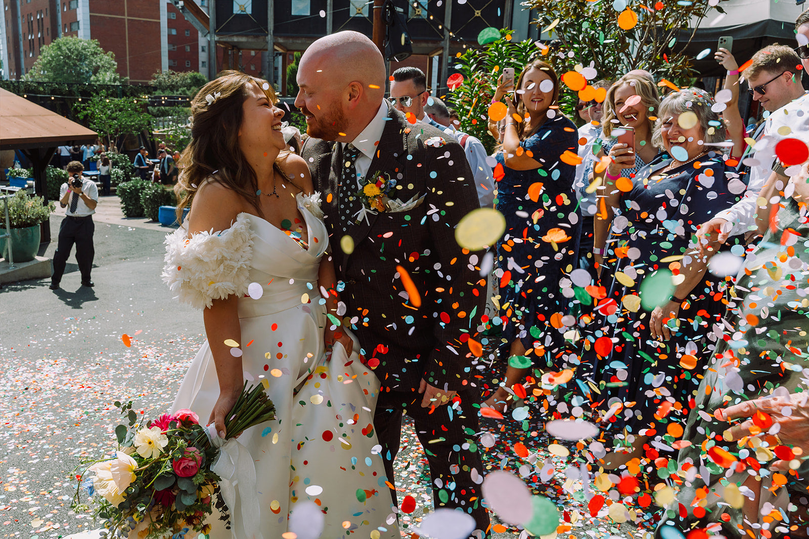 A bride in an off-the-shoulder white wedding gown and a groom in a dark patterned suit are surrounded by guests throwing colorful confetti in an outdoor street setting.