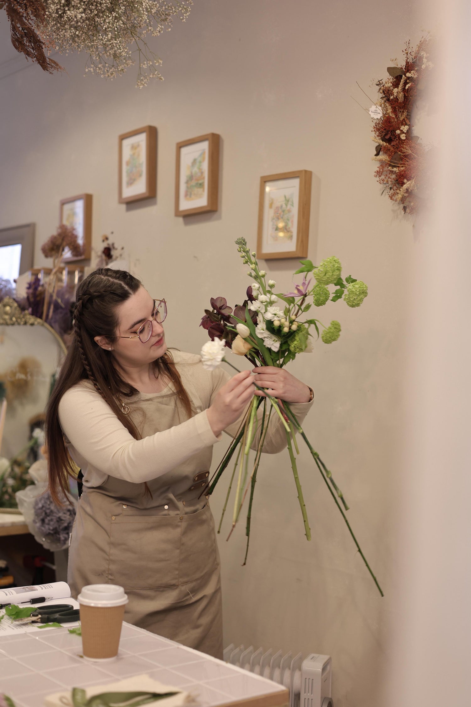Jordan, owner of Blooming Edens, arranging a bridal bouquet in a floral shop with framed pictures on the wall.