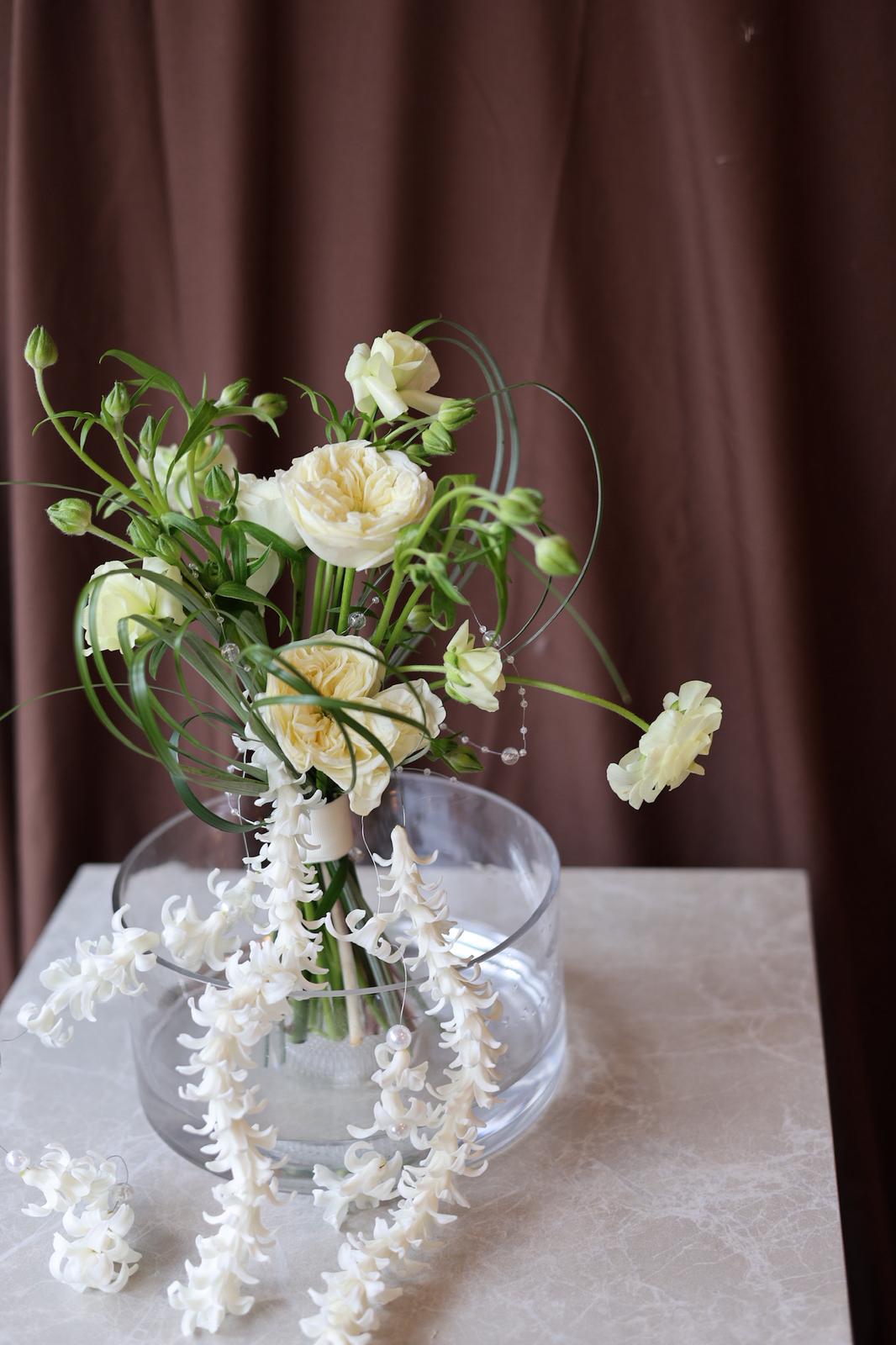 Bouquet of white flowers in a clear vase on a marble surface with a brown curtain background